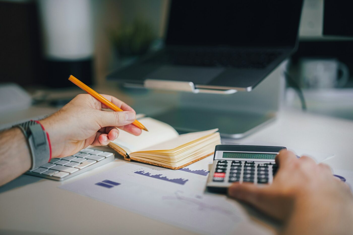 Zwei Hände am Schreibtisch: die eine hält einen Bleistift über einem aufgeschlagenen Notizbuch, die andere tippt auf einem Taschenrechner – daneben Tastatur, Laptop und ein ausgedrucktes Diagramm