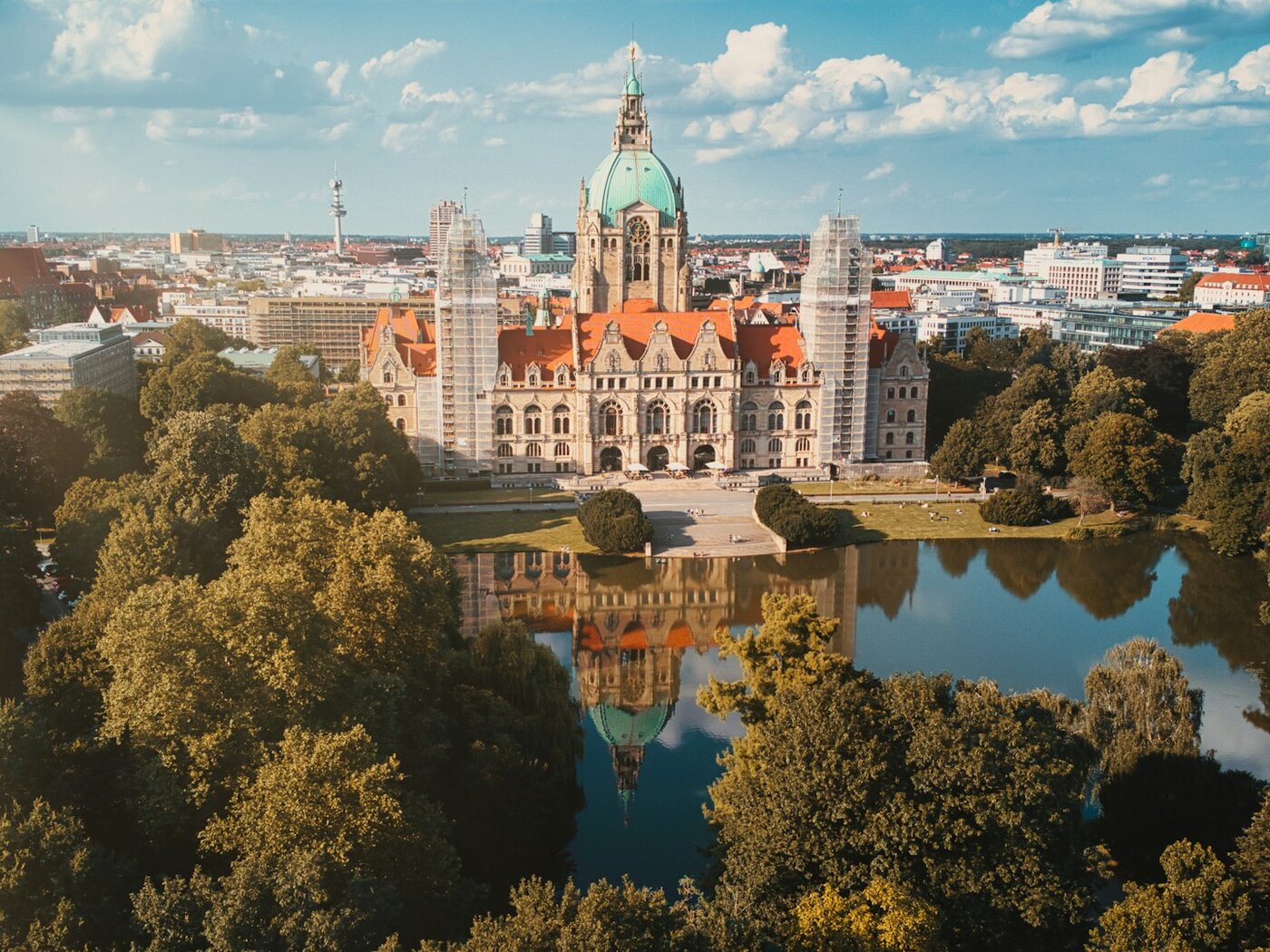 Luftbild des Neuen Rathauses in Hannover mit grüner Kuppel und rotem Dach, gespiegelt im Maschteich und umrahmt von den Bäumen des Maschparks, dahinter die Stadtsilhouette