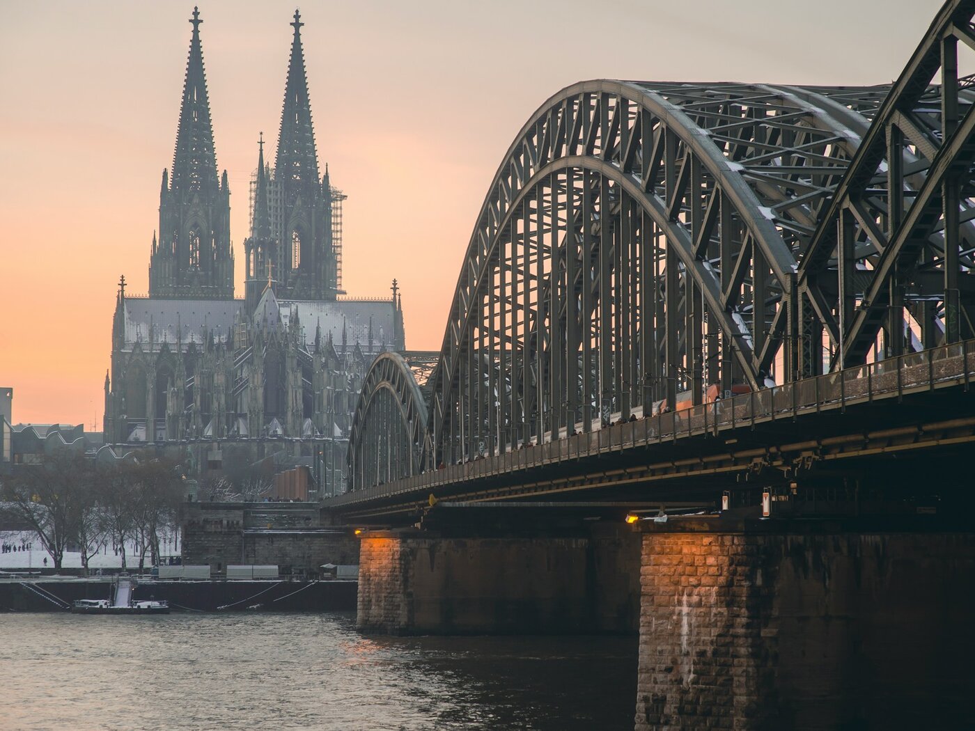 Der Kölner Dom mit seinen beiden Türmen im Morgenlicht, davor die Stahlbögen der Hohenzollernbrücke über dem Rhein, im Vordergrund verschneite Uferbäume