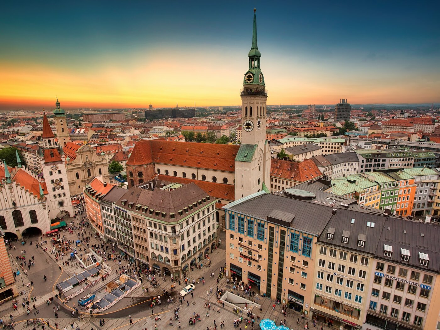 Blick über den Münchner Marienplatz im Abendlicht: der Turm der Peterskirche mit Uhr und grüner Haube, links das Alte Rathaus, dahinter die roten Dächer der Altstadt
