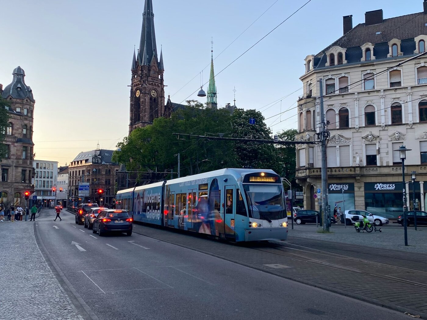 Blaue Saarbahn am Saarbrücker Rathausplatz in St. Johann in der Abenddämmerung, dahinter der Turm der neugotischen Johanneskirche und Gründerzeithäuser